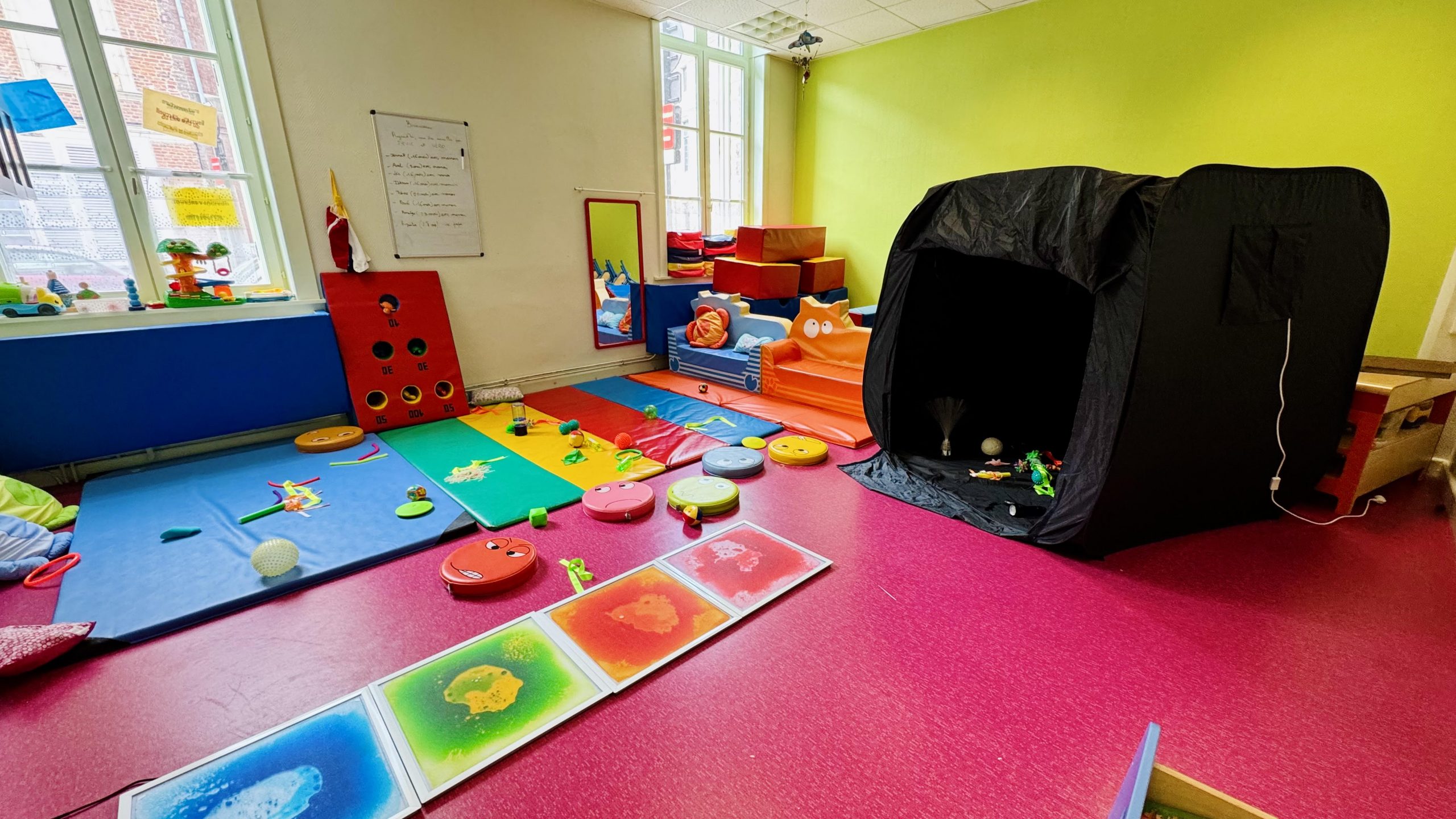 Salle du LAEP Les p'tits Loupiots à Douai avec tapis de motricité colorés, jouets d'éveil au sol, panneaux lumineux sensoriels, modules de psychomotricité et tente noire d'exploration, dans un espace lumineux aux murs vert anisSalle du LAEP Les p'tits Loupiots à Douai avec tapis de motricité colorés, jouets d'éveil au sol, panneaux lumineux sensoriels, modules de psychomotricité et tente noire d'exploration, dans un espace lumineux aux murs vert anis.