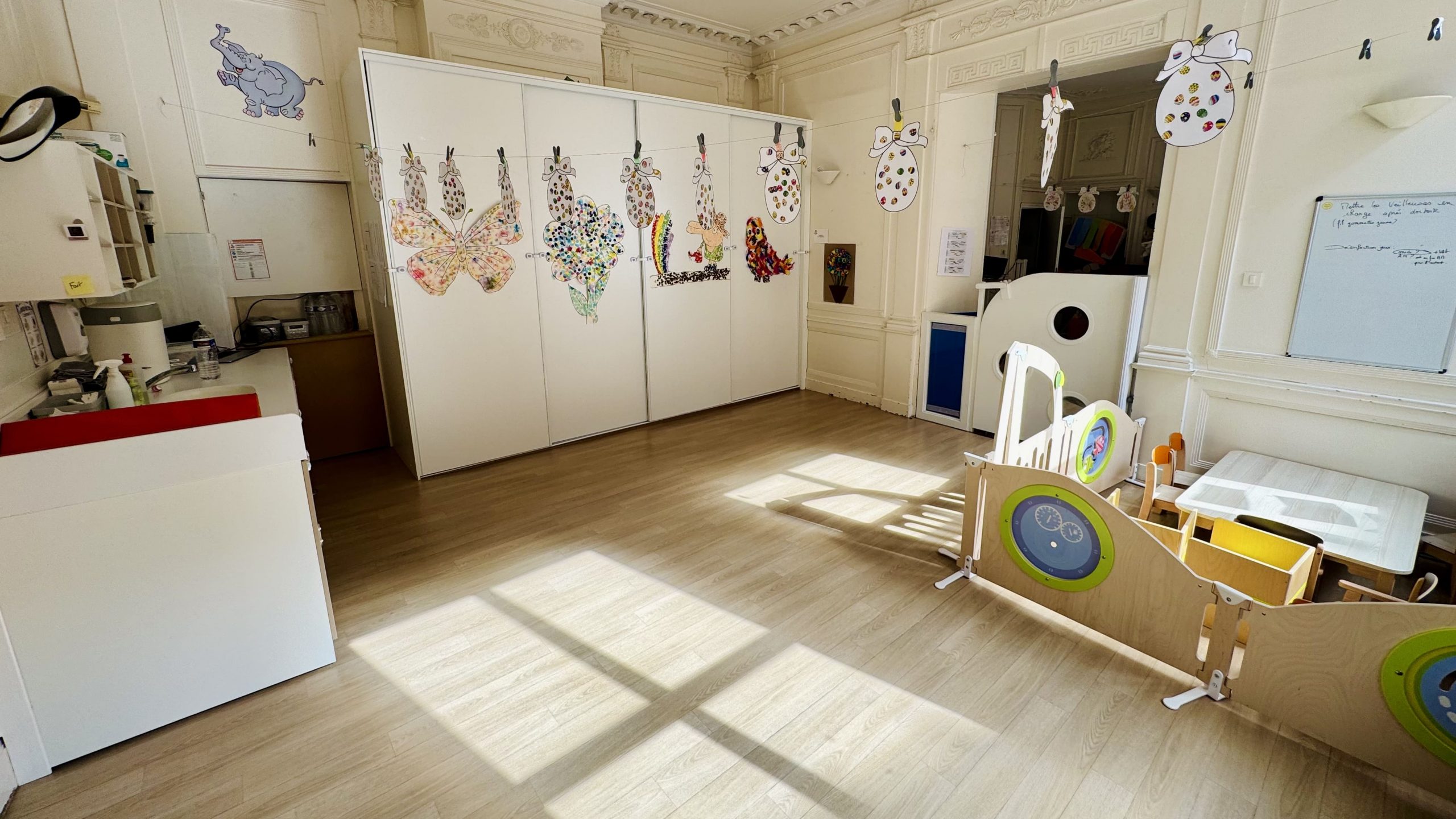 Salle de la section Oursons avec parquet clair, lumière naturelle marquée au sol, créations d'enfants affichées sur portes de placard et matériel de motricité au premier plan.