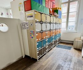 Dès l'entrée, chaque enfant a son propre espace personnel — son casier, son repère à lui. Espace d'accueil de la crèche Petit-à-Petit avec rangée de casiers colorés individuels, hublot décoratif et créations d'enfants suspendues au plafond