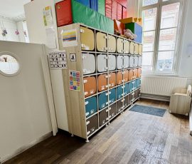 Dès l'entrée, chaque enfant a son propre espace personnel — son casier, son repère à lui. Espace d'accueil de la crèche Petit-à-Petit avec rangée de casiers colorés individuels, hublot décoratif et créations d'enfants suspendues au plafond