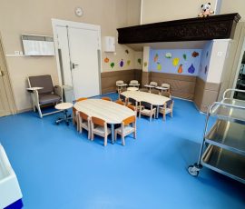 L'heure des repas : des tables conviviales, des chaises à leur taille, et des légumes qui donnent des idées ! Salle à manger de la crèche Petit-à-Petit avec tables ovales et chaises adaptées aux tout-petits, sol bleu vif, frise de légumes colorés peints sur le mur et accès direct à la cuisine