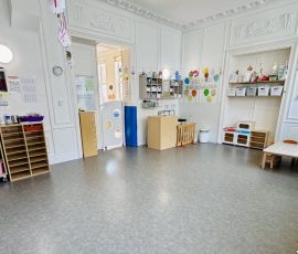 La section des Loupiots : un espace de vie à leur mesure, dans un cadre chaleureux et structuré. Salle principale de la section Loupiots avec tables et chaises adaptées aux tout-petits, rangements colorés étiquetés, coin symbolique et décoration murale dans une pièce à hauts plafonds ornementés.