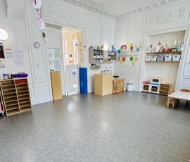 La section des Loupiots : un espace de vie à leur mesure, dans un cadre chaleureux et structuré. Salle principale de la section Loupiots avec tables et chaises adaptées aux tout-petits, rangements colorés étiquetés, coin symbolique et décoration murale dans une pièce à hauts plafonds ornementés.