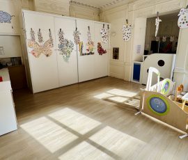 La section des Oursons baigne dans la lumière : un espace ouvert pour bouger, explorer et créer. Salle de la section Oursons avec parquet clair, lumière naturelle marquée au sol, créations d'enfants affichées sur portes de placard et matériel de motricité au premier plan.
