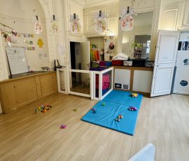 La section des Poussins : un espace douillet et sécurisé pour les tout-tous-petits, avec tapis de jeu, jouets d'éveil et plafonds chargés d'histoires. Section des Poussins de la crèche Petit-à-Petit avec grand tapis bleu au sol, jouets d'éveil colorés, parc bébé, décoration de Pâques suspendue au plafond et moulures ornementales d'un bâtiment historique de Douai.